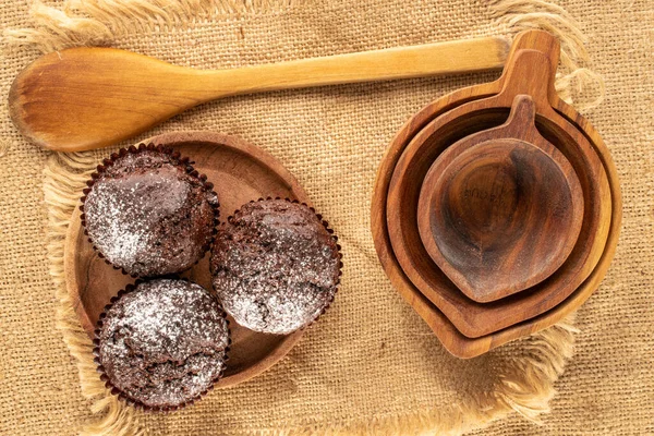 Three homemade chocolate cupcakes with wooden saucer, three cups, wooden spoon and jute napkin on jute cloth, macro, top view.