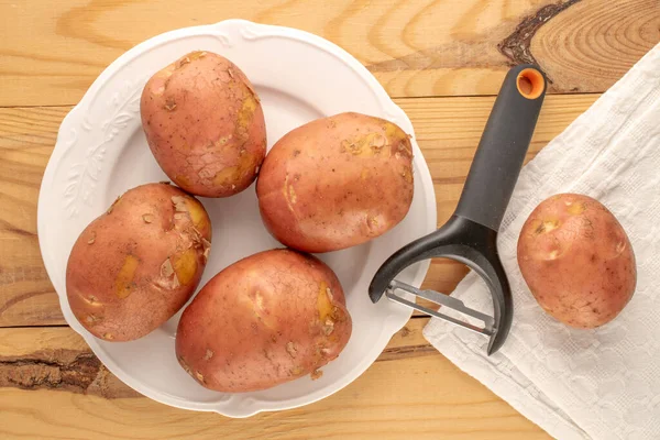 几个生的粉红土豆 有一个白色的陶瓷盘 一个白色的亚麻布餐巾和一把用来剥皮蔬菜的刀 宏观的 放在一张木制桌子上 顶部看 — 图库照片