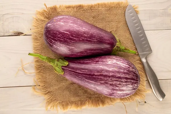 Two ripe eggplants with jute napkin and knife on wooden table, macro, top view.