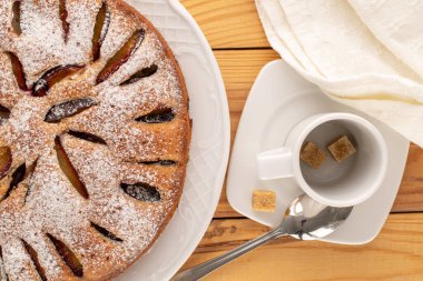 Beyaz seramik çanaklı ev yapımı erikli kek, fincan, çay tabağı, bıçak ve çatal ahşap masa, makro, üst manzara.