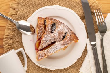 Beyaz seramik çanaklı ev yapımı erikli kek, fincan, çay tabağı, bıçak ve çatal ahşap masa, makro, üst manzara.