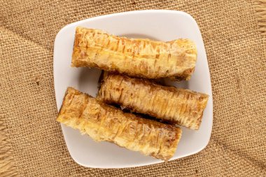 Üç parça tatlı baklava, beyaz seramik bir fincan tabağı jüt kumaşın üzerinde, yakın plan, üst manzara..