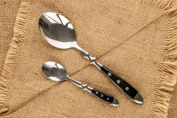 Two metal spoons on a jute napkin, macro, top view.