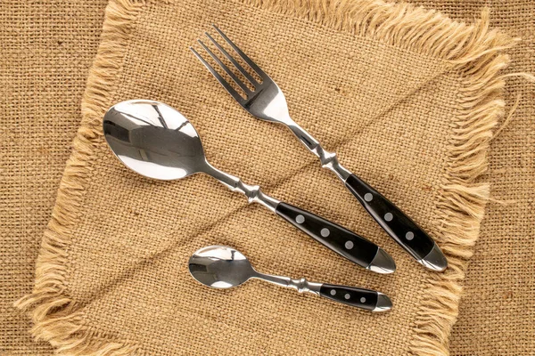 Two metal spoons and fork on a jute napkin, macro, top view.