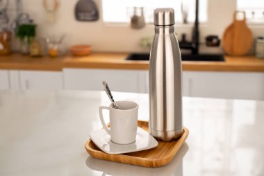 Bir metal su ya da elektrikli süpürge yalıtımlı kahve şişesi, ahşap tepside ve mutfak masasında beyaz bardak..