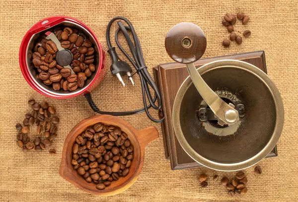 One manual wooden coffee grinder, one electric coffee grinder with coffee beans and a wooden cup with coffee beans on a jute cloth, macro, top view.