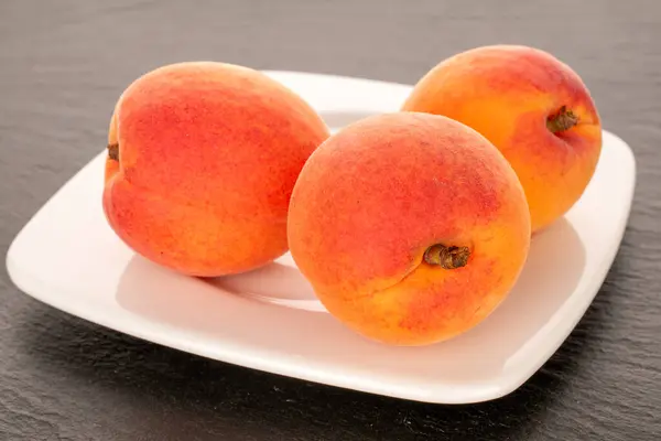 stock image Three juicy apricots on a white ceramic plate on slate stone, macro.