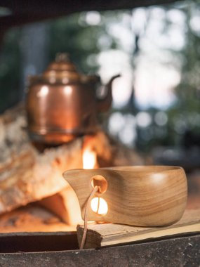 Ön planda odaklanmış bir kamp ahşap kupası. Arka plandaki yangında antika bir bakır çaydanlık ısınıyor..