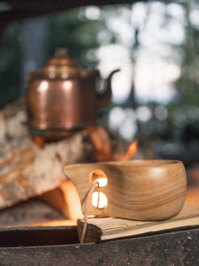 Ön planda odaklanmış bir kamp ahşap kupası. Arka plandaki yangında antika bir bakır çaydanlık ısınıyor..