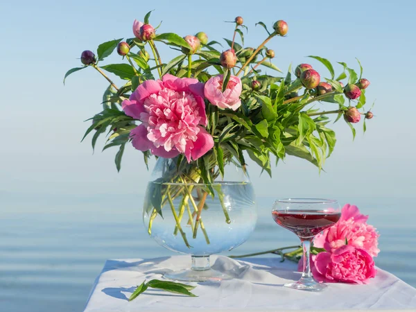 A bouquet of pink peonies in a glass vase and a glass of red wine on a table covered with a white tablecloth, against the background of the lake.
