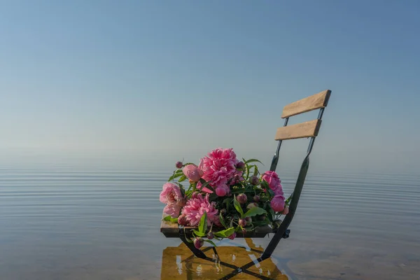 A wooden chair with a bouquet of gorgeous pink peonies stands in the center surrounded by lake water. A gentle and relaxing composition.