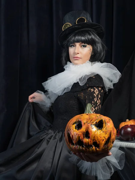 A beautiful brunette in a vintage black dress, a white frill and a vintage hat holds a scary pumpkin in her hands. The concept of a happy Halloween holiday.