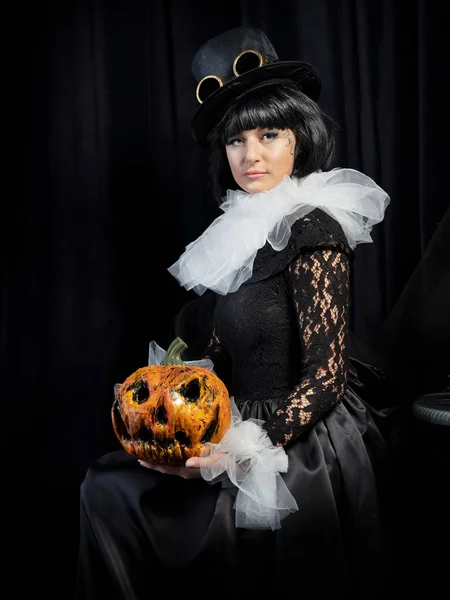 A beautiful brunette in a vintage black dress, a white frill and a vintage hat holds a scary pumpkin in her hands. The concept of a happy Halloween holiday.