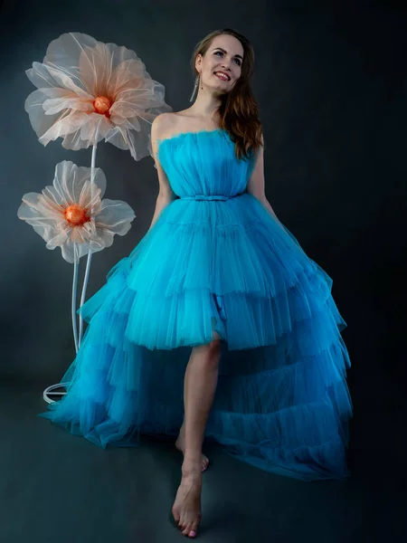 stock image Portrait of a beautiful young woman with long hair in a lush blue tulle dress in the studio against a background of decorative flowers. 