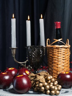 Dekoratif bir hayat yanan mumlar, hasır bir şarap şişesi, kırmızı elmalar ve koyu mavi arka planda altın bir şarap bahçesi..