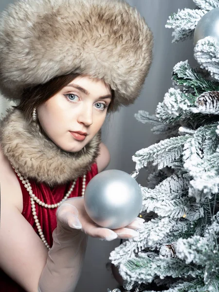 A beautiful woman in a fur hat, fur collar and pearls at the Christmas tree. Russian style. Close-up.