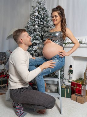 Bir adam hamile karısının önünde oturuyor ve bir Noel ağacı ve şöminenin arka planına gülümsüyor. Gelecekteki güzel ebeveynlerin portresi. Mutlu yıllar ve mutlu Noeller..