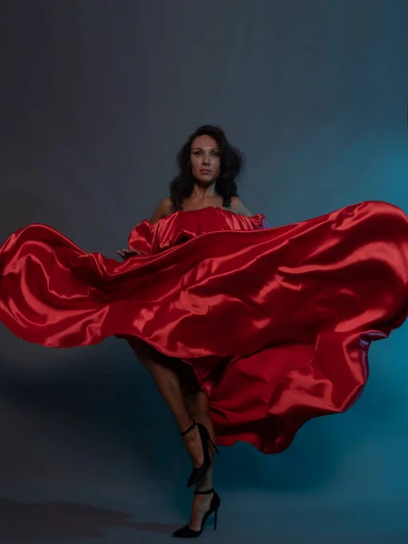 A beautiful brunette in a red flying dress poses in the studio on a gray background.