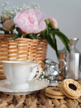 Hasır sepette pembe şakayıklı sanatsal bir hayat, bir fincan kahve, krema ve kurabiye. Dikey fotoğraf.