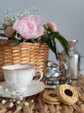 Hasır sepette pembe şakayıklı sanatsal bir hayat, bir fincan kahve, krema ve kurabiye. Dikey fotoğraf.