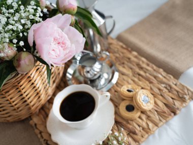 Hasır sepette pembe şakayıklı sanatsal bir hayat, bir fincan kahve, krema ve kurabiye. Üst Manzara. Yatay fotoğraf.