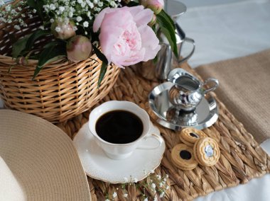 Hasır sepette pembe şakayıklı sanatsal bir hayat, bir fincan kahve, krema ve kurabiye. Üst Manzara. Yatay fotoğraf.