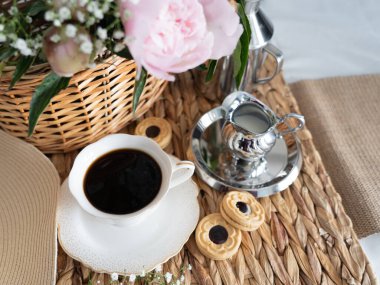 Hasır sepette pembe şakayıklı sanatsal bir hayat, bir fincan kahve, krema ve kurabiye. Üst Manzara. Yatay fotoğraf.