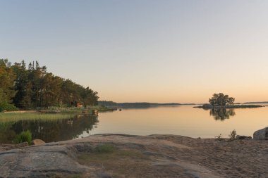 Nagu, Finlandiya - 12.06.2022: Gün batımından sonra sakin deniz kıyısı. Bir sıra ağaç ve ufukta küçük bir ada. Ön planda Rocky Coast var. Finlandiya doğası. Doğal arka plan. Metin için boşluk.