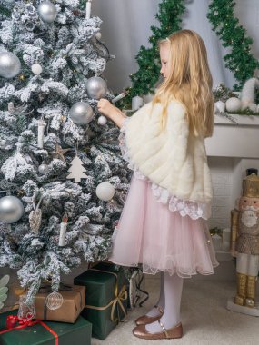 Pembe elbiseli ve kürklü uzun sarı saçlı küçük bir kızın portresi. Bir kız Noel ağacı oyuncaklarına hayran. Tam boy portre. Sihirli Noel zamanı.