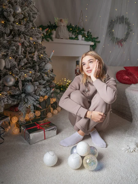 A young blonde girl with a short haircut in a cozy suit is dreaming about the Christmas tree in the background of the fireplace.