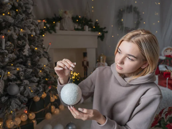 A young blonde girl with a short haircut in cozy clothes hangs Christmas balls on a Christmas tree on the background of the fireplace.
