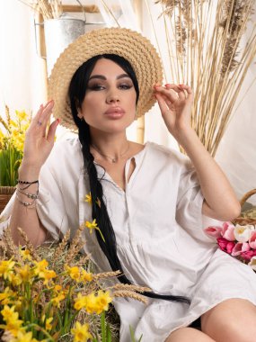 Güzel bir esmer, uzun örgülü, beyaz bir elbise ve hasır bir şapka giyiyor. Stüdyoda saman ve nergis çiçekleri üzerinde fotoğraf bölgesi.
