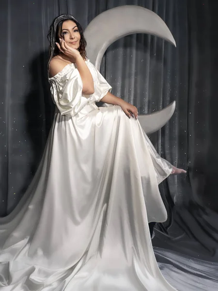Portrait of a woman from the back in a white satin dress with a long train. She is against the background of a large decorative moon.