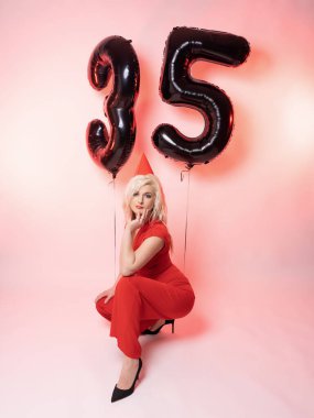 Kırmızı gece elbisesi ve 35 numaralı balonlu güzel sarışın. Doğum gününü kutluyor. Pembe arkaplan.
