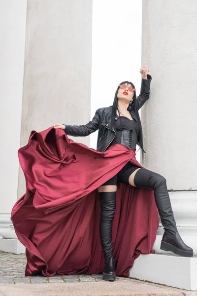 Woman with long hair, perfect figure, sexy. she is in a satin, long red skirt, leather jackets. Posing in the city at the tall ancient columns