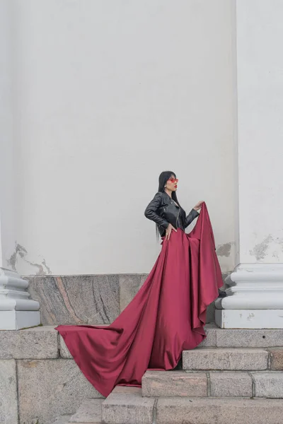 Woman with long hair, perfect figure, sexy. she is in a satin, long red skirt, leather jackets. Posing in the city at the tall ancient columns