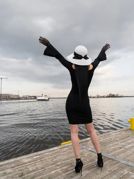 A woman with a perfect figure in a short black dress, on the pier in a white hat. Meets the ferry, waving his hands