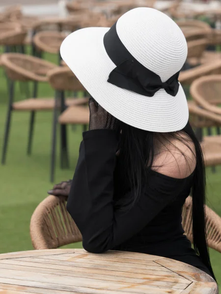 A woman in a black dress, at a table in a summer cafe in a white hat. One