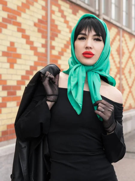 A woman with a perfect figure and red lips in a short black dress, a bright scarf on her head. Posing against the background of a brick wall