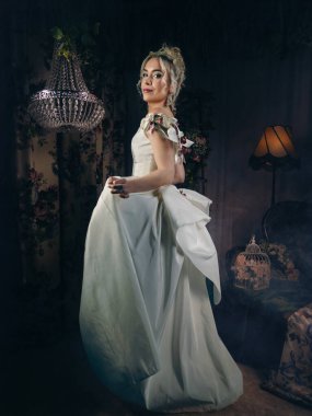 Viktorya dönemi romantizmi tarzında lüks bir stüdyo fotoğrafı. Zarif bir elbise içindeki çekici bir kadın şehvetli çiçeklerle bezenmiş bir iç mekanda antika mobilyalar, retro ışıklandırma ve eski mankenler gibi antika elementlerle poz veriyor. 