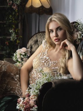 Lüks işlemeli bir gece elbisesi içinde zarif bir sarışın çiçeklerle çevrili eski bir iç mekânda poz veriyor. Büyüleyici bir görünüş, sofistike, aristokratik bir cazibe ve romantik bir atmosfer..