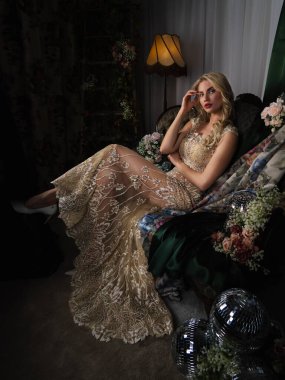 Lüks işlemeli bir gece elbisesi içinde zarif bir sarışın çiçeklerle çevrili eski bir iç mekânda poz veriyor. Büyüleyici bir görünüş, sofistike, aristokratik bir cazibe ve romantik bir atmosfer..