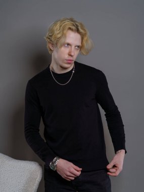 Beyaz dalgalı saçlı, siyah kazak giyen, gri arka planda bir sandalyeye oturan genç bir adam. Basit ama şık kompozisyonu onun rahat ve kendinden emin tavrını vurguluyor..