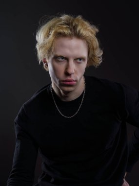 Siyah elbiseli, dalgalı sarı saçlı, koyu bir arka planı olan genç bir adamın portresi. Yüzünde ciddi bir ifade ve bakışları çok yoğun. Dramatik ışıklandırma yüz hatlarını ve saçlarını ön plana çıkarıyor.