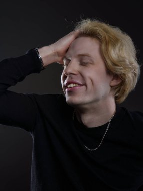 Siyah elbiseli, dalgalı sarı saçlı, koyu bir arka planı olan genç bir adamın portresi. O neşeli bir ifadeyle gülüyor. Atmosferik aydınlatma yüz hatlarını ve saç dokusunu vurguluyor.