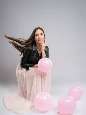 Model testinde genç bir kadın, tam boy. Hareket, dalgalı pembe elbise, deri ceketli kontrast. Tarafsız arkaplan, pembe balonlar neşe ve hafiflik ekler