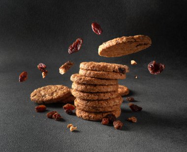 Kuru üzümlü yulaflı kurabiyeler koyu gri arka planda bir yığının içine düşüyor.