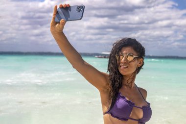 Selfie çekerken bikinili seksi kız cennet sahilinin tadını çıkarıyor. Turist seyahatinden ve cep telefonuyla fotoğraf çeken tropikal bir plajdan hoşlanıyor. Bikinili kız..