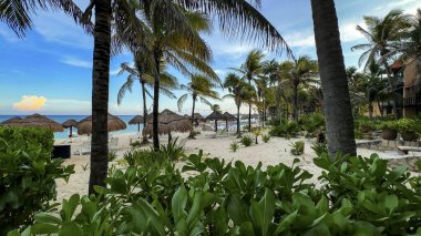 Arka planda kristal berrak turkuaz su ve yemyeşil Karayip bitki örtüsü ile, güzel, beyaz kumlu bir plajda lüks bir tatil köyünün binalarında yürümek.