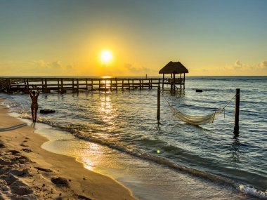 Meksika Karayipleri 'ndeki güzel bir kumsalda turuncu bir gün doğumunda yoga yapan seksi bir kadın. Arkasında ahşap iskele ve palapa var. Kristal berrak turkuaz su üzerinde..